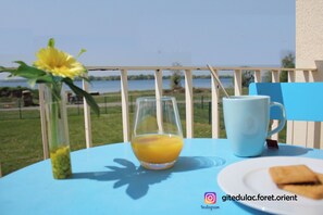 Ein Tisch mit einem Orangensaftglas, einer weißen Tasse und einem Teller mit Gebäck, draußen an einem blauen Tisch mit Seeblick und weißem Zaun.