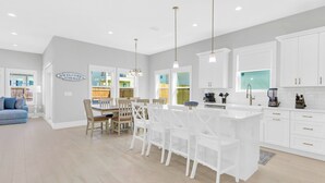 A modern kitchen with white cabinetry, a central island with bar stools, and a dining area with a wooden table and chairs. - Tailwinds: Free golf cart, Private hot tub, Pool, Game room, 4min to beach (Miramar Beach)
