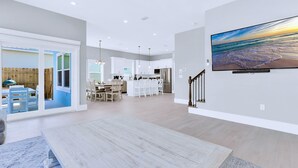 A spacious living area with a large TV, a dining table, and a kitchen in the background. - Tailwinds: Free golf cart, Private hot tub, Pool, Game room, 4min to beach (Miramar Beach)