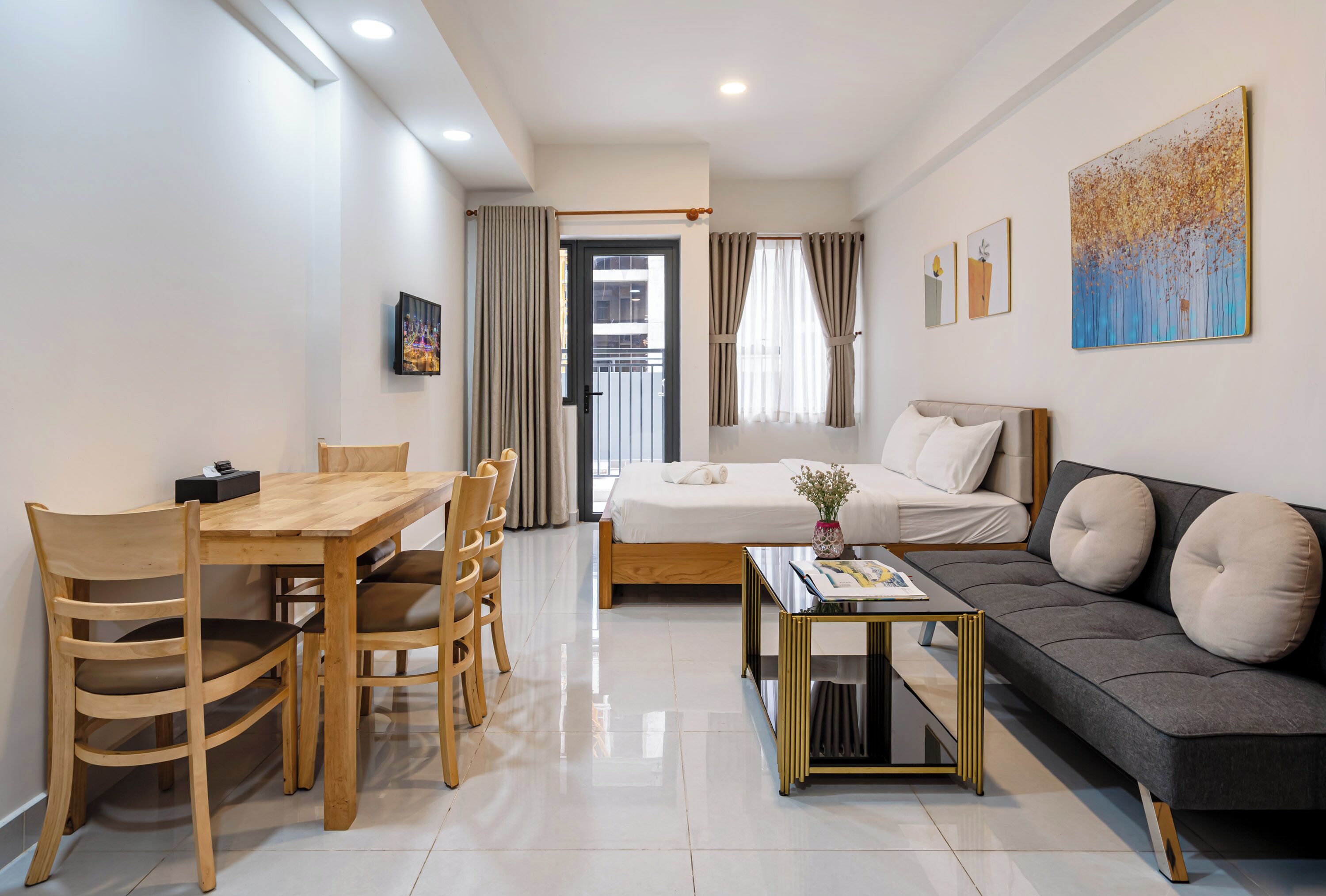 A modern hotel room with a bed, sofa, dining table, chairs, and artwork on the walls.
