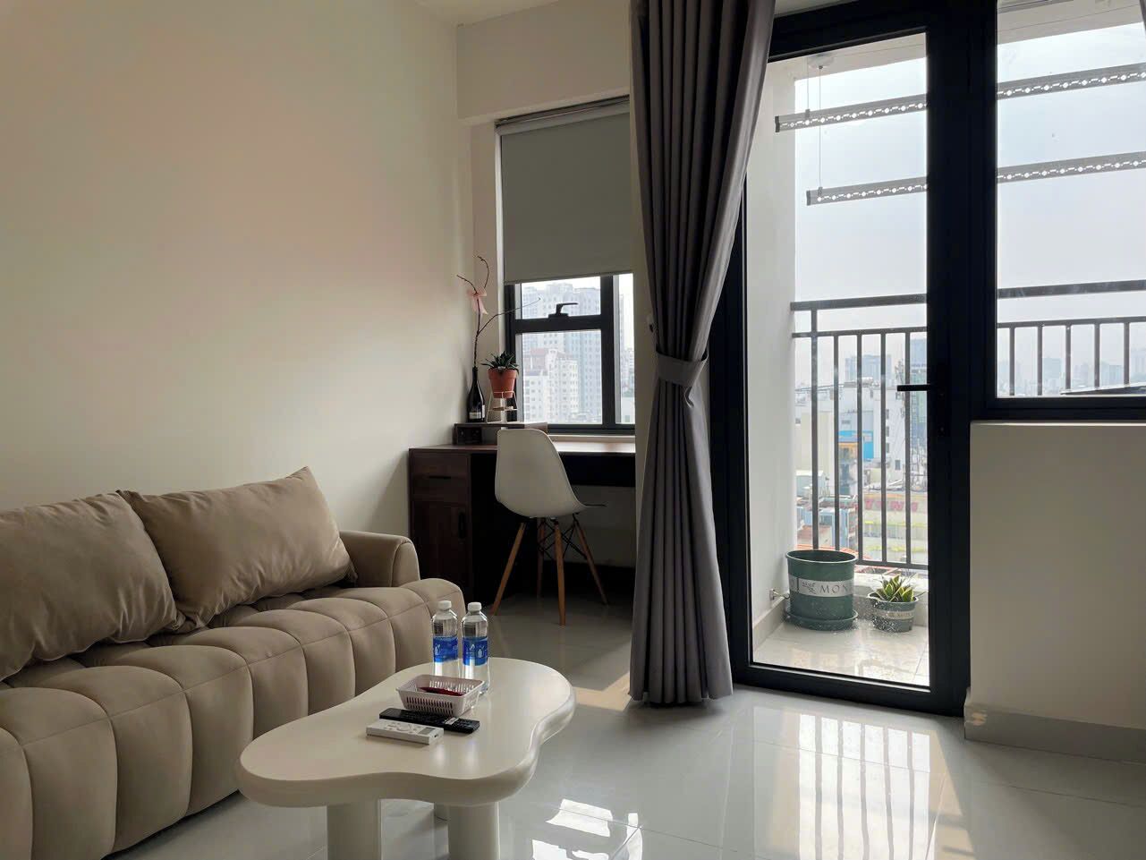 En moderne stue med sofa, sofabord og skrivebord. Der er et vindue med gardiner, en balkon med gelænder, og udsigt til bygninger.