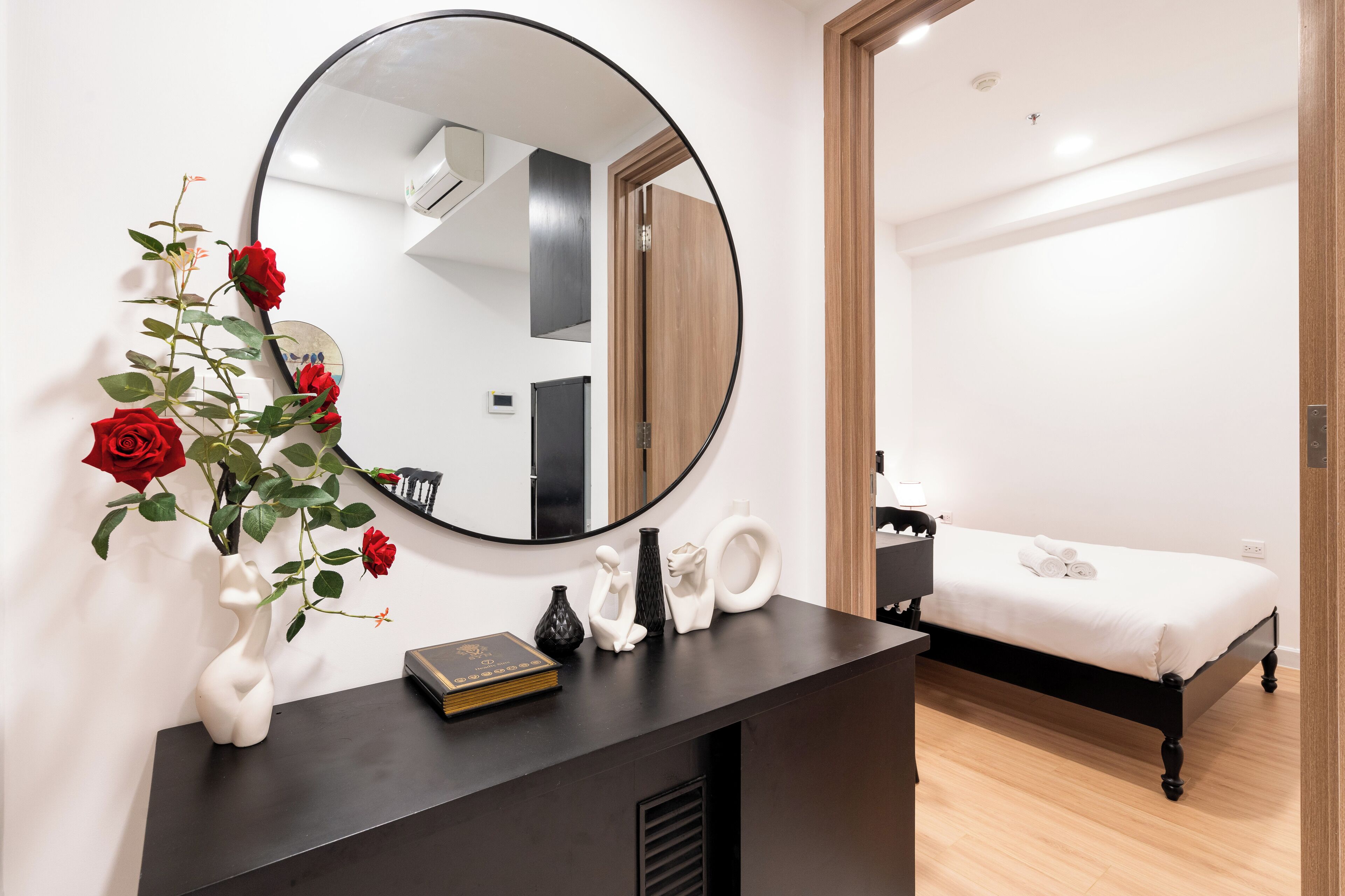 A modern bedroom with a large round mirror, a bedside table with a book, a vase with red roses, and a neatly made bed.