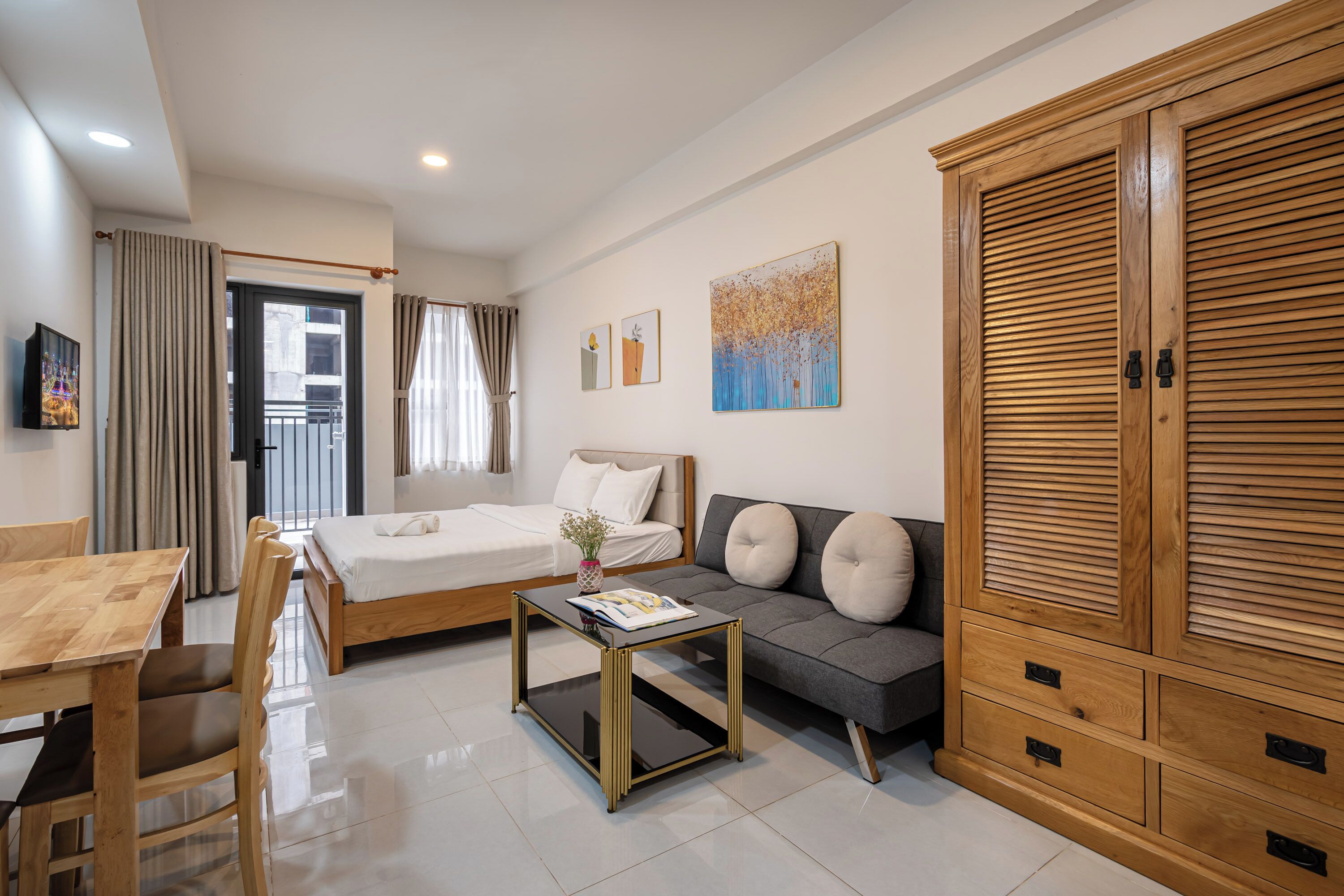 A modern hotel room with a bed, sofa, wooden wardrobe, and a dining area.