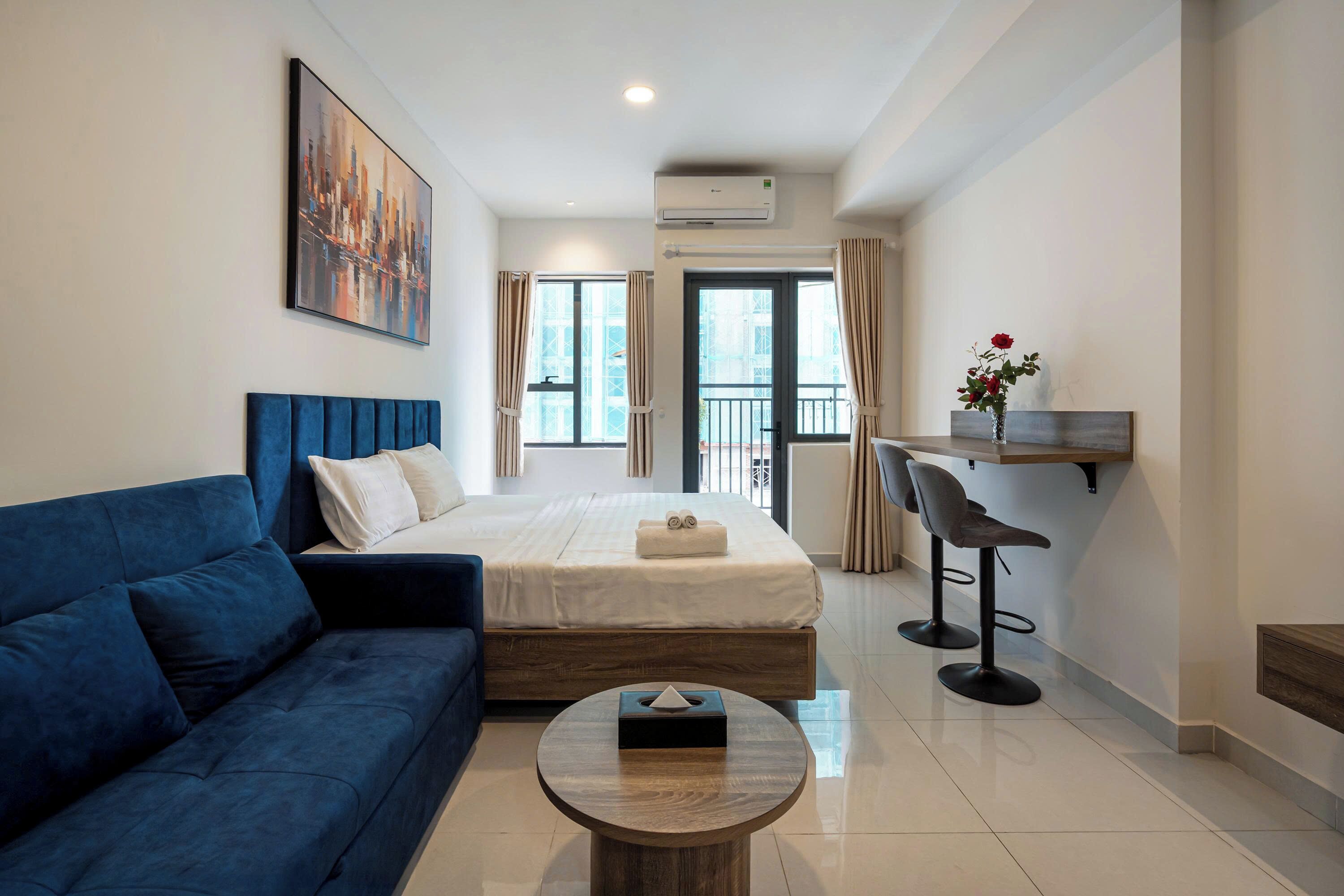 A modern hotel room with a large bed, a blue sofa, a round coffee table, and a desk with two chairs.