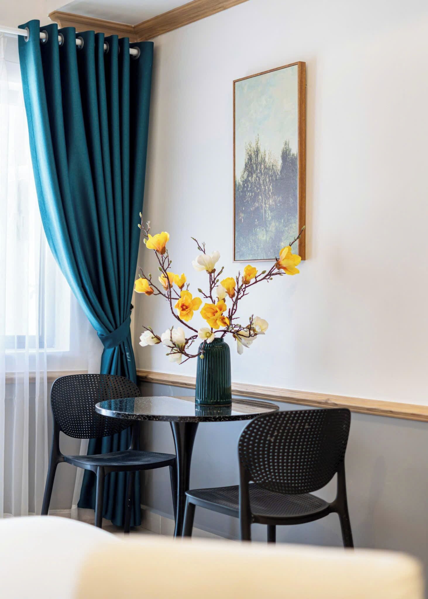 A dining area with a round table, two chairs, a vase with yellow flowers, teal curtains, and a framed picture on the wall.