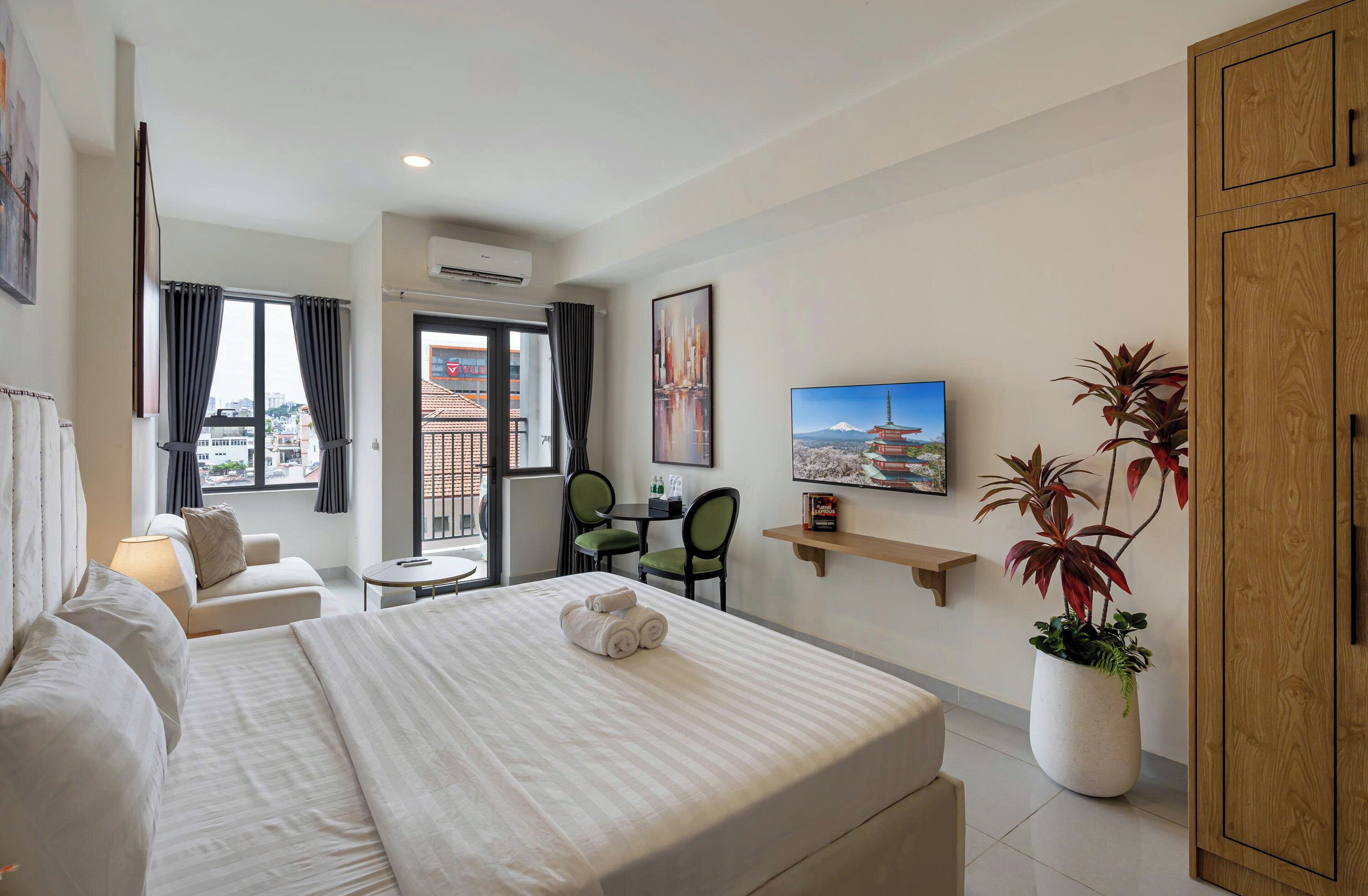 A hotel room with a bed, a desk, a chair, a small table, a TV, a potted plant, and a view of the city.