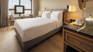 Habitación de hotel con una cama grande, un escritorio, una silla, un televisor, un baño con espejo y vistas a la ciudad desde la ventana.