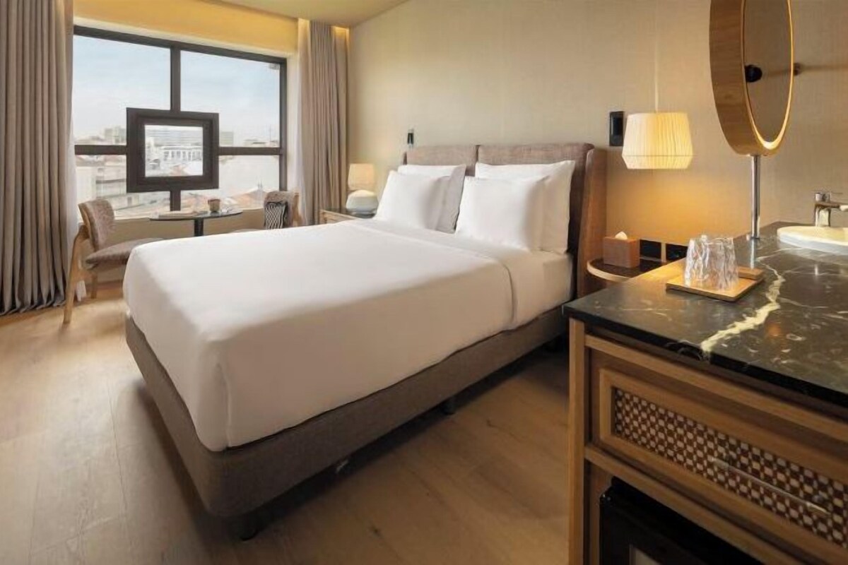 Een hotelkamer met een groot bed, een bureau, een stoel, een televisie, een badkamer met een spiegel en uitzicht op de stad vanuit het raam.