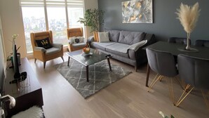 A modern living room with a grey sofa, a coffee table, and a dining table with chairs. There is a large window with blinds, a plant, and a painting on the wall. - MERDZ Spa & Fitness Sky Resident  (Skopje)