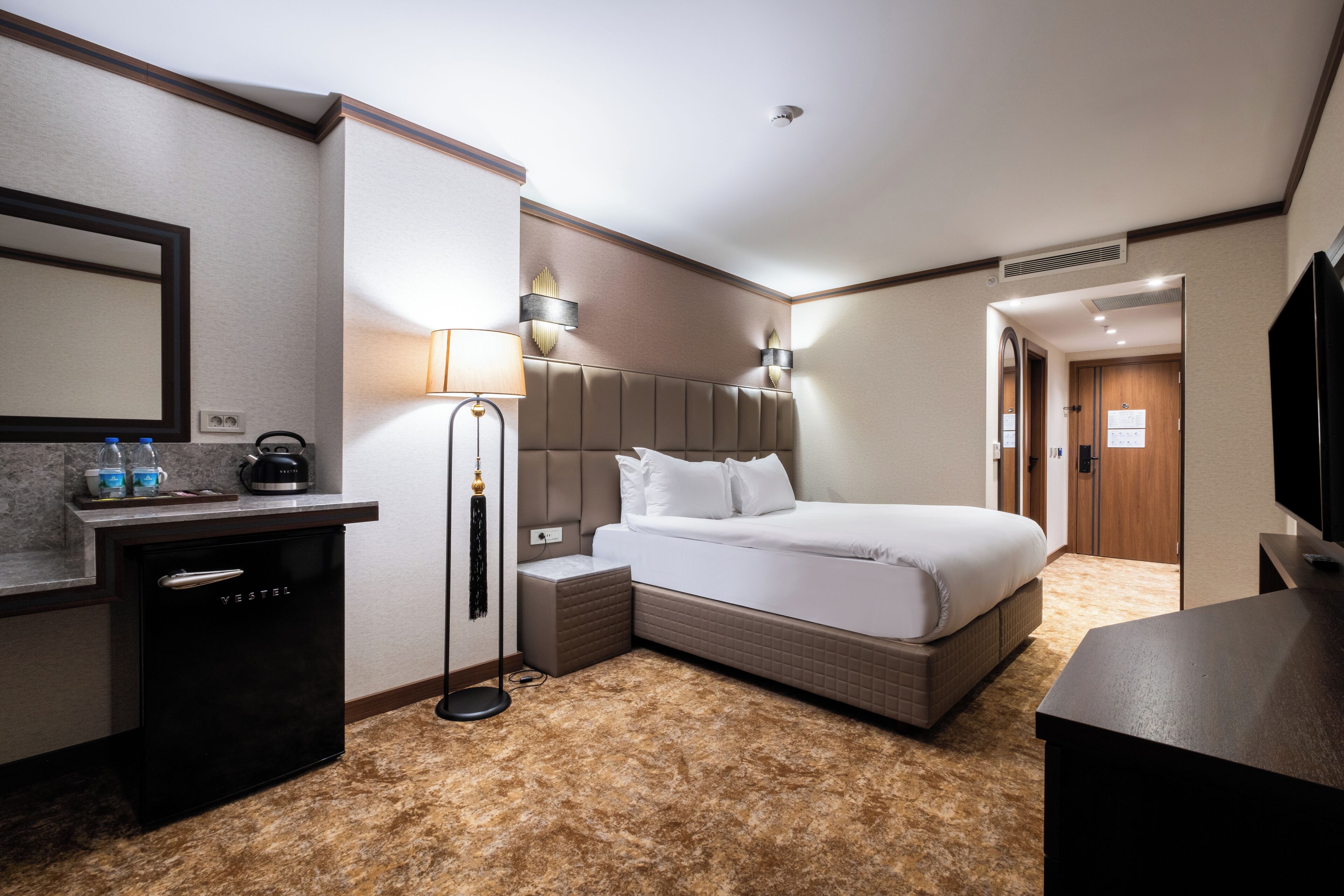 Habitación de hotel con cama, mesita de noche, lámpara y un minibar.