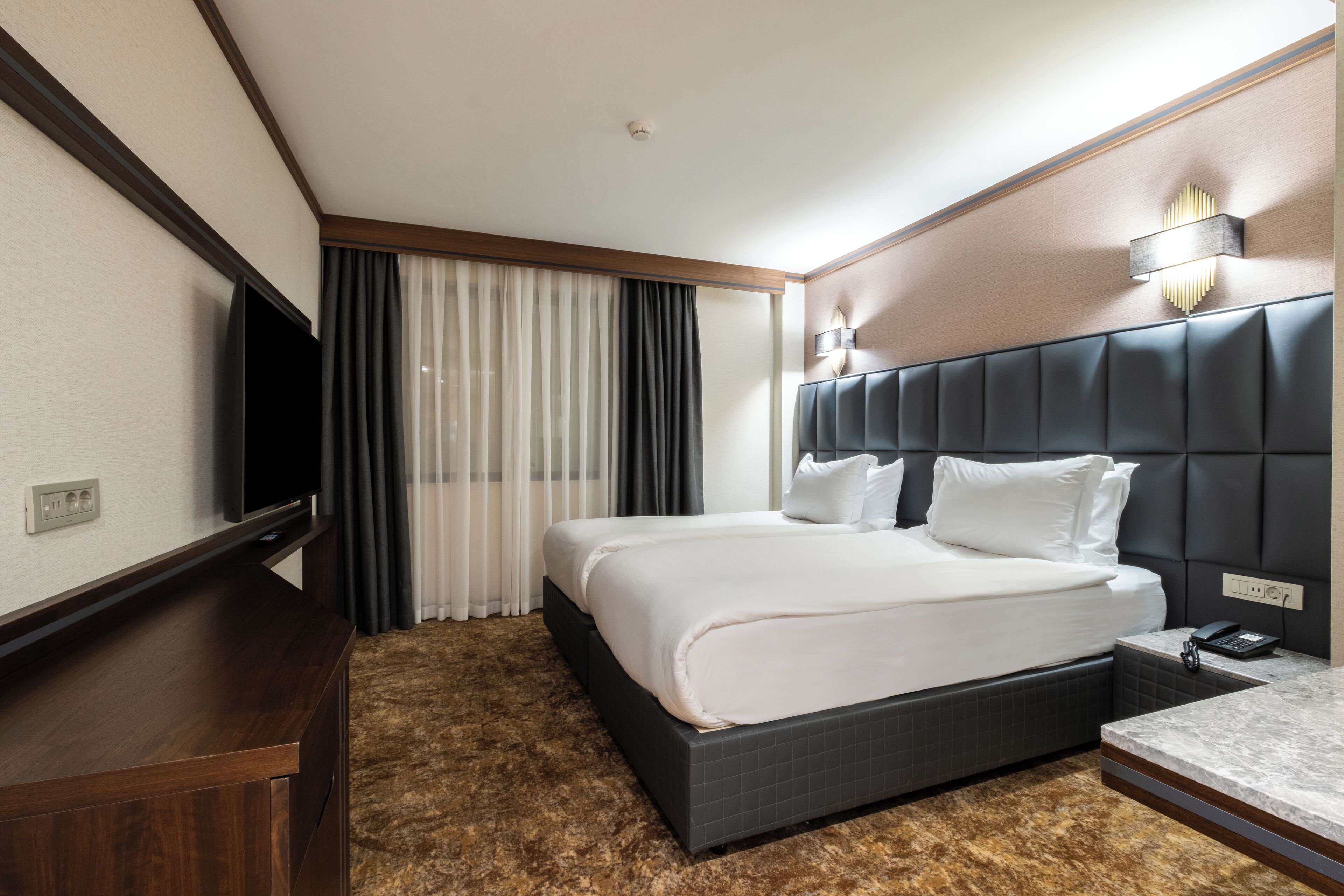 A hotel room with a large bed, a flat-screen TV, a bedside table with a lamp, and a window with curtains.