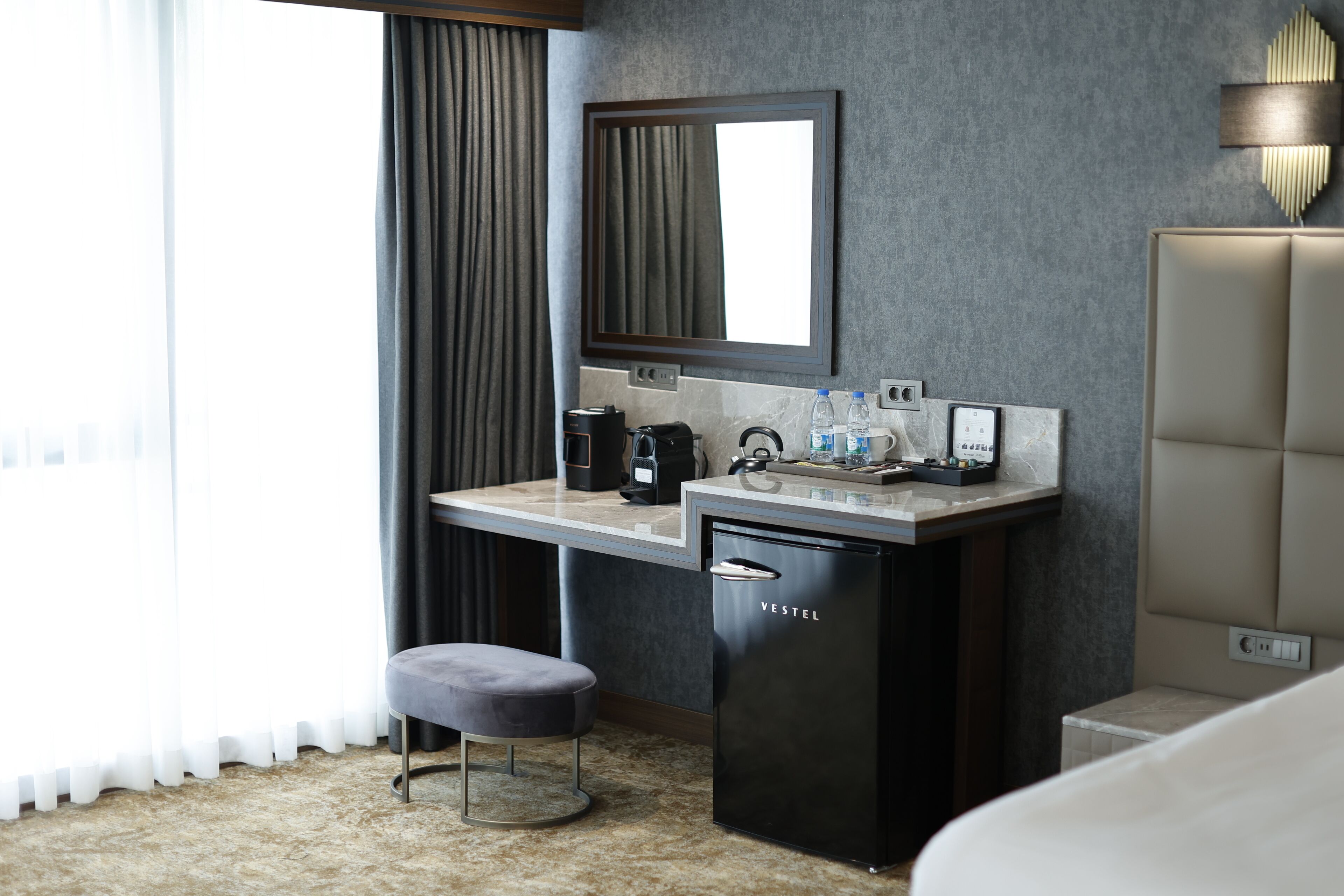 Une chambre d’hôtel équipée d’un bureau, d’un minibar, d’un petit tabouret, d’un lit, d’un grand miroir et d’une fenêtre avec des rideaux.