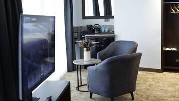 Una habitación de hotel moderna con un televisor de pantalla plana grande, un sillón cómodo, una pequeña mesa redonda con un jarrón de flores y una kitchenette al fondo.