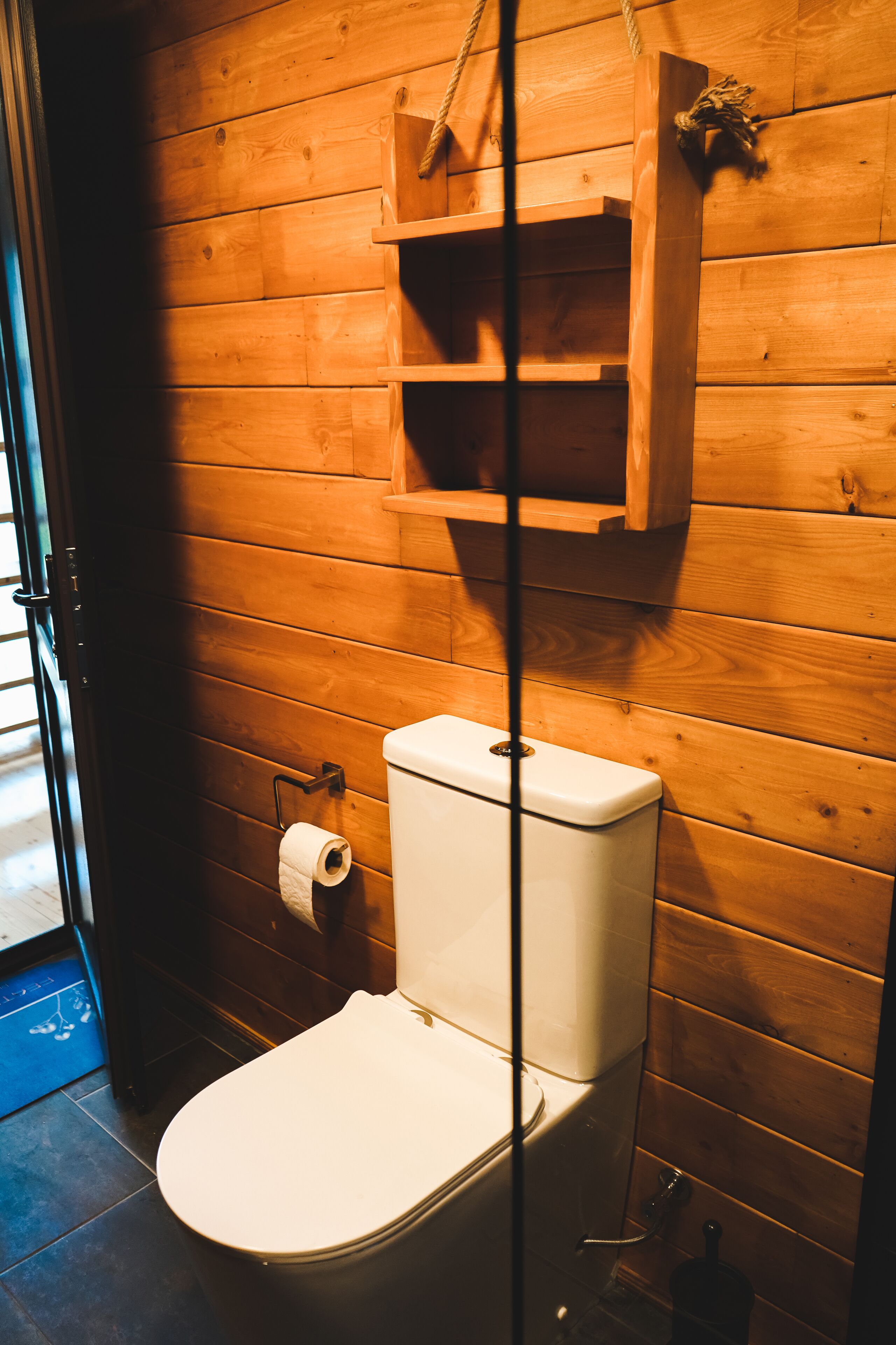 A wooden-paneled bathroom with a toilet, a shelf above the toilet, and a hanging toilet paper holder.