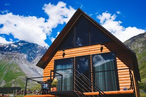 A wooden log cabin with large windows and a balcony, set against a mountainous backdrop. - Traveler's Cottage in Kazbegi (Kazbegi)