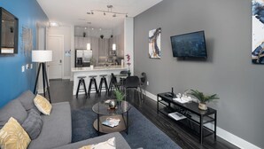 A modern living room with a grey sofa, a coffee table, and a TV. There is a kitchen in the background with a dining area. - Luxury Serviced Apartment in Uptown CLT (Charlotte)