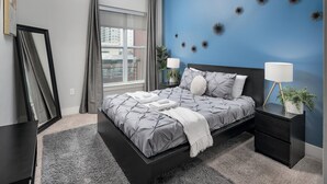 A modern bedroom with a large bed, two bedside tables, a nightstand, a window with curtains, and decorative wall art. - Luxury Serviced Apartment in Uptown CLT (Charlotte)