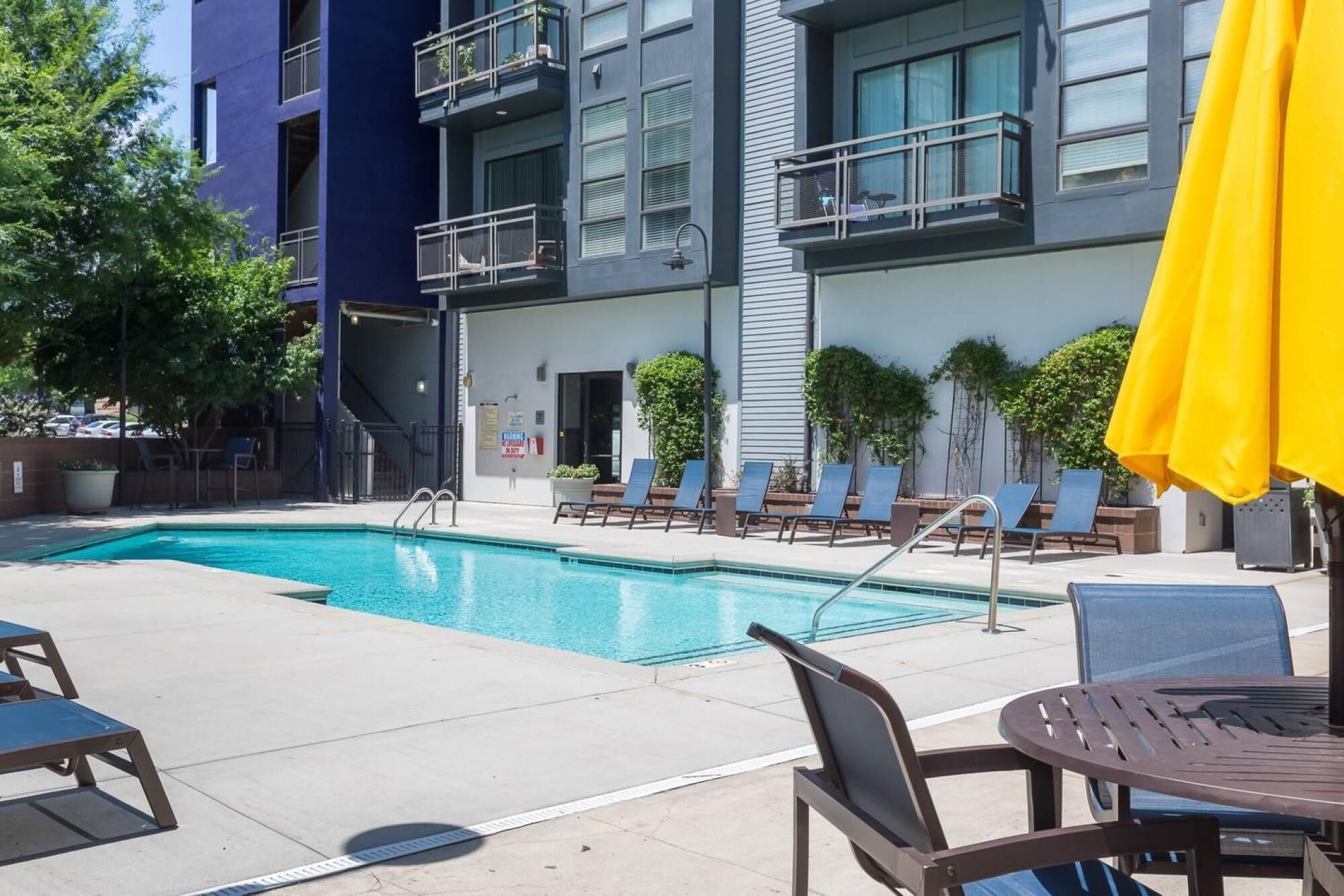 Un espace piscine aménagé avec des chaises longues et une table, entouré d’un bâtiment doté de balcons et de fenêtres.
