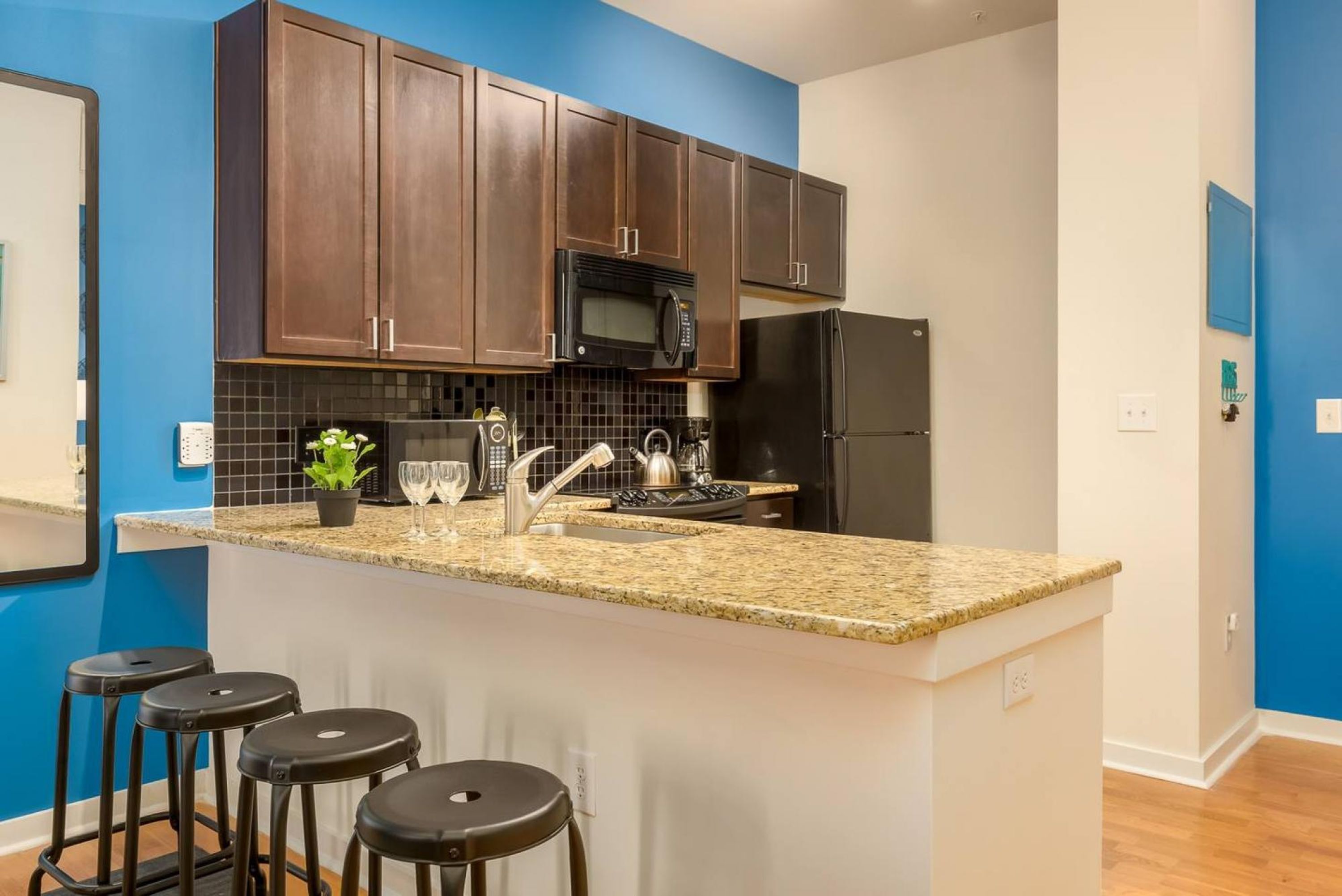 Una cocina con armarios de madera oscura, encimera de granito, pileta de acero inoxidable y refrigerador negro, además de un microondas. Hay tres taburetes negros en la isla.
