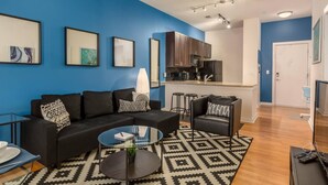 Un appartement moderne avec un mur d’accent bleu, un canapé noir, une table basse en verre, un coin cuisine et un téléviseur.