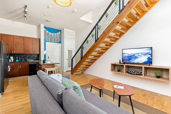 Un apartamento moderno con una escalera de madera, una cocina, una sala de estar con sofá y televisor, y una pequeña mesa de comedor.