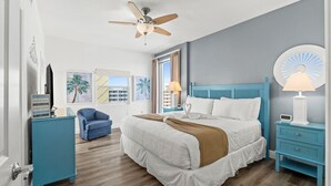 A bedroom with a bed, a ceiling fan, a nightstand, a lamp, a chair, and a window with curtains. - PI 613: Salty Sunsets - Beachfront Bliss with Unmatched Ocean Views (Fort Walton Beach)