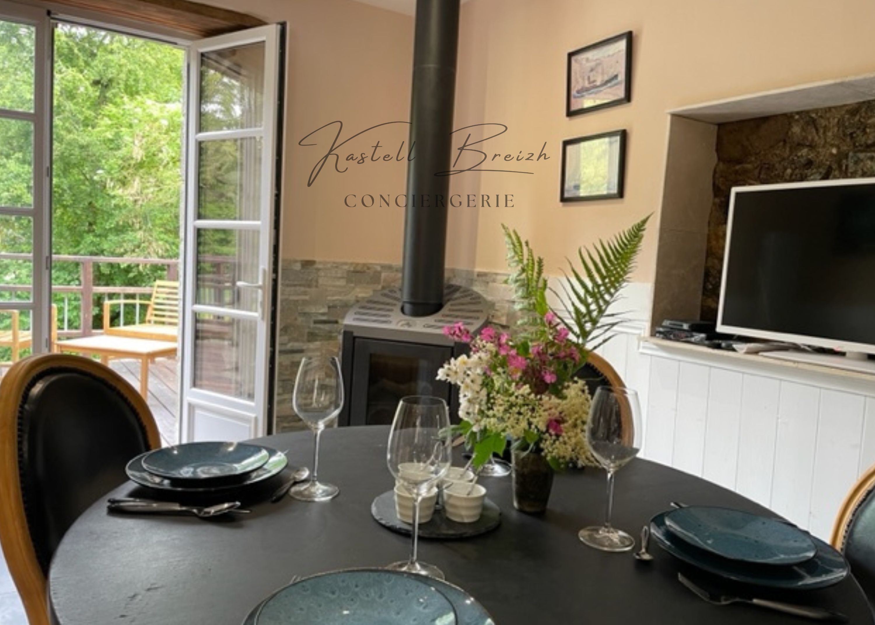 A dining table set with plates, glasses, and a vase with flowers, with a fireplace and TV in the background.
