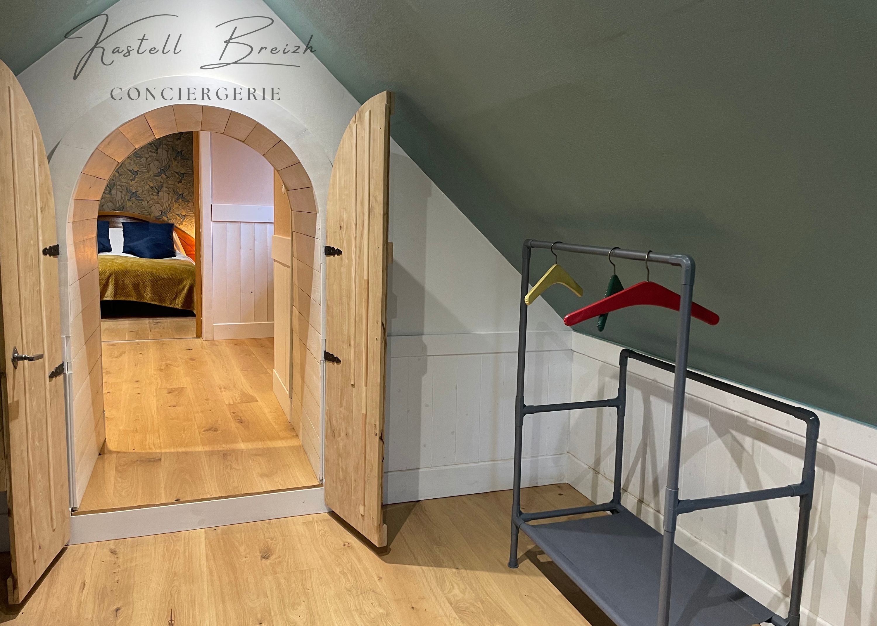 Une pièce avec un plancher en bois, un lit recouvert d’une couverture verte, une armoire en bois et un portant à vêtements avec des cintres.
