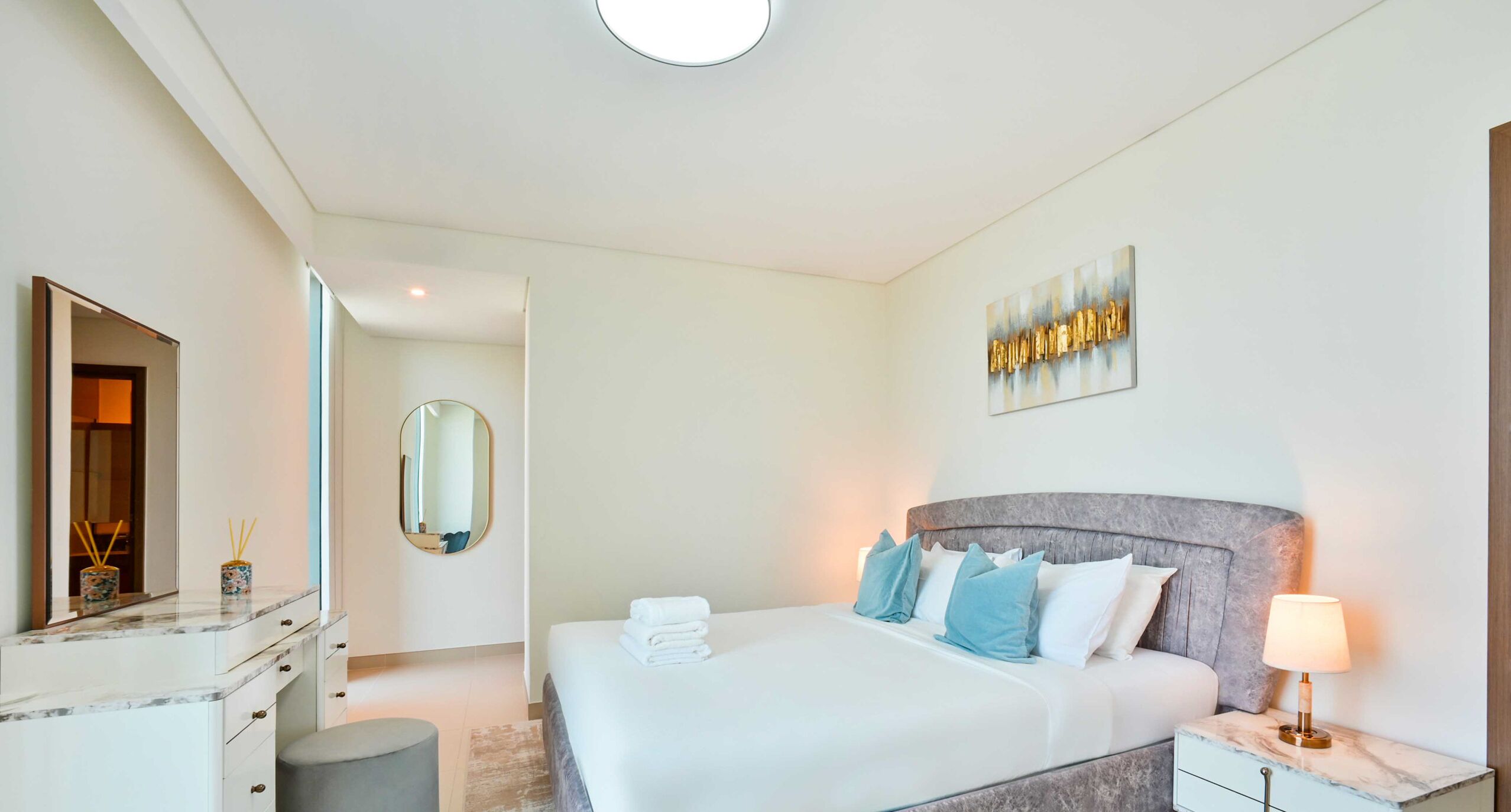 Une chambre d’hôtel moderne avec un grand lit, une table de chevet avec une lampe, une commode, un miroir et une œuvre d’art murale.