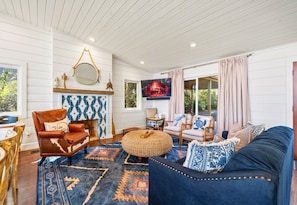 A cozy living room with a leather armchair, a blue sofa, a woven ottoman, and a television mounted on the wall.