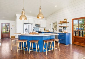 A kitchen with a central island, wooden stools, pendant lighting, and a view of the outdoors.