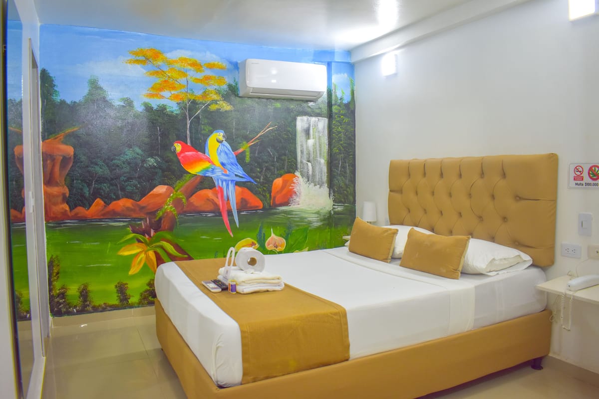 Habitación de hotel con un mural de paisaje tropical, una cama con cabecera capitonada y una mesita de noche con teléfono.