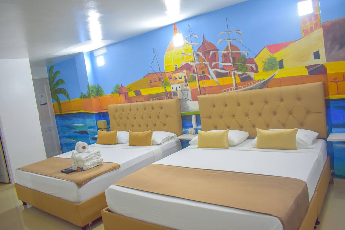 Habitación de hotel con dos camas, cada una con un cabecero capitonado, y un mural de una escena costera en la pared.