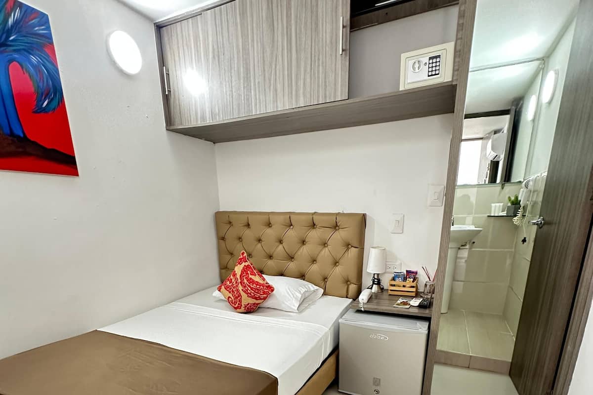 Una habitación de hotel con cama, mesita de noche, una pequeña mesa, un espejo y un baño visible a través de una puerta abierta.
