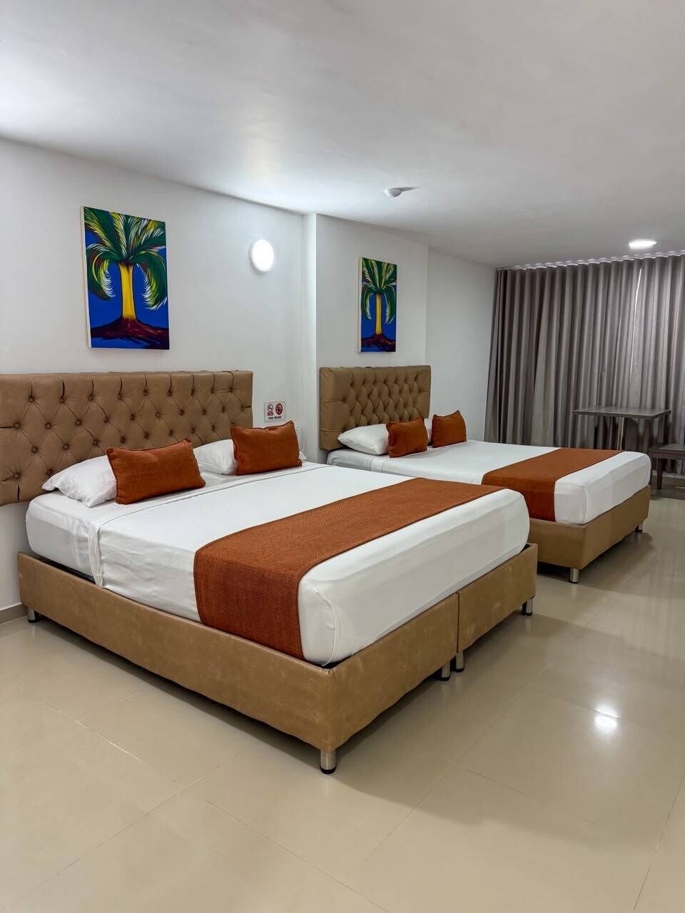A hotel room with two beds, a large window with curtains, and two paintings on the wall.
