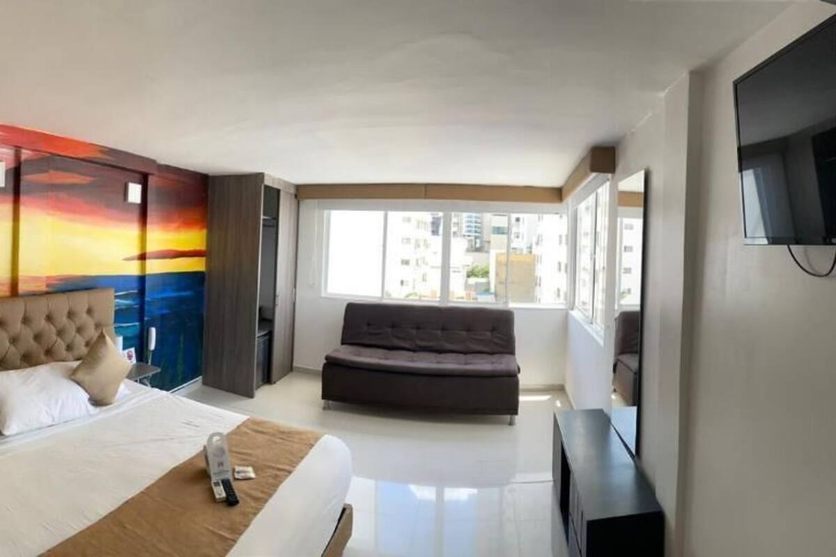 Habitación de hotel con una cama grande, un sofá, un televisor y un mural de un atardecer sobre el océano.