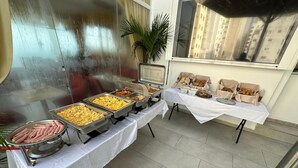 A buffet setup with various food items in metal containers, a potted plant, and a window with a view of buildings. - Hotel Hernandez CTG (Cartagena)