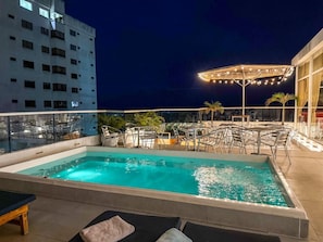 A rooftop pool with clear blue water, surrounded by metal chairs and tables, and a covered area with string lights.