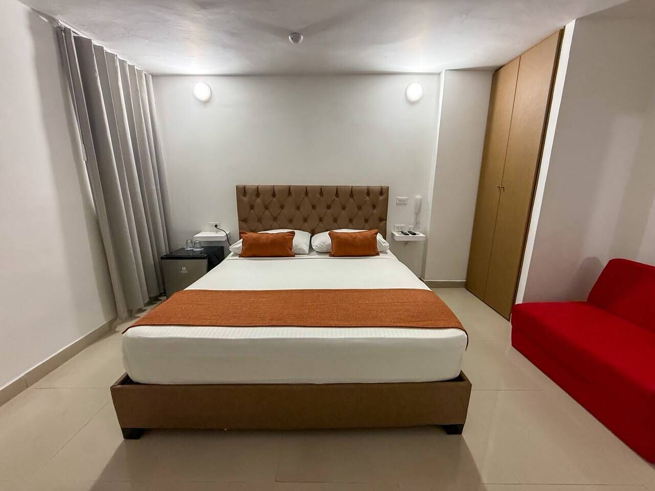 A hotel room with a large bed, a bedside table, a nightstand, a red sofa, and a wooden wardrobe.