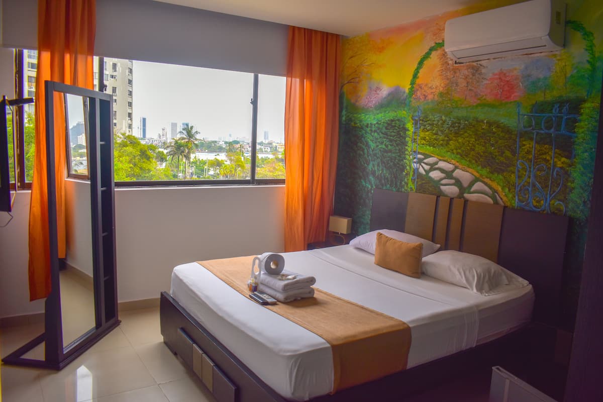 Una habitación de hotel con cama, ventana con cortinas, un cuadro en la pared y un equipo de aire acondicionado.