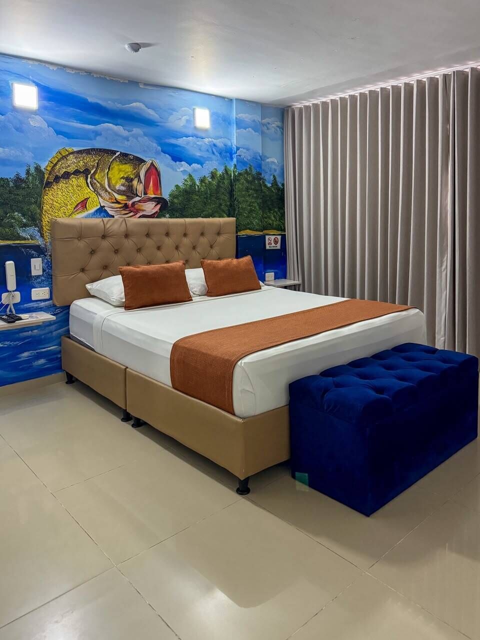 A bedroom with a bed, a headboard, a nightstand, a blue ottoman, and a mural on the wall.