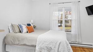 A bedroom with a bed, pillows, a bedside table, a lamp, a window with curtains, and a TV mounted on the wall. - Studio 1 ~ Lovely Apartment, Downtown! (Moncton)
