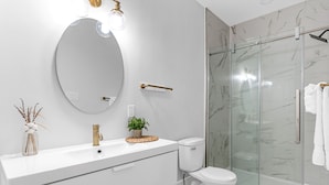 A modern bathroom with a white vanity, a round mirror, a gold faucet, and a glass shower enclosure. - Studio 1 ~ Lovely Apartment, Downtown! (Moncton)