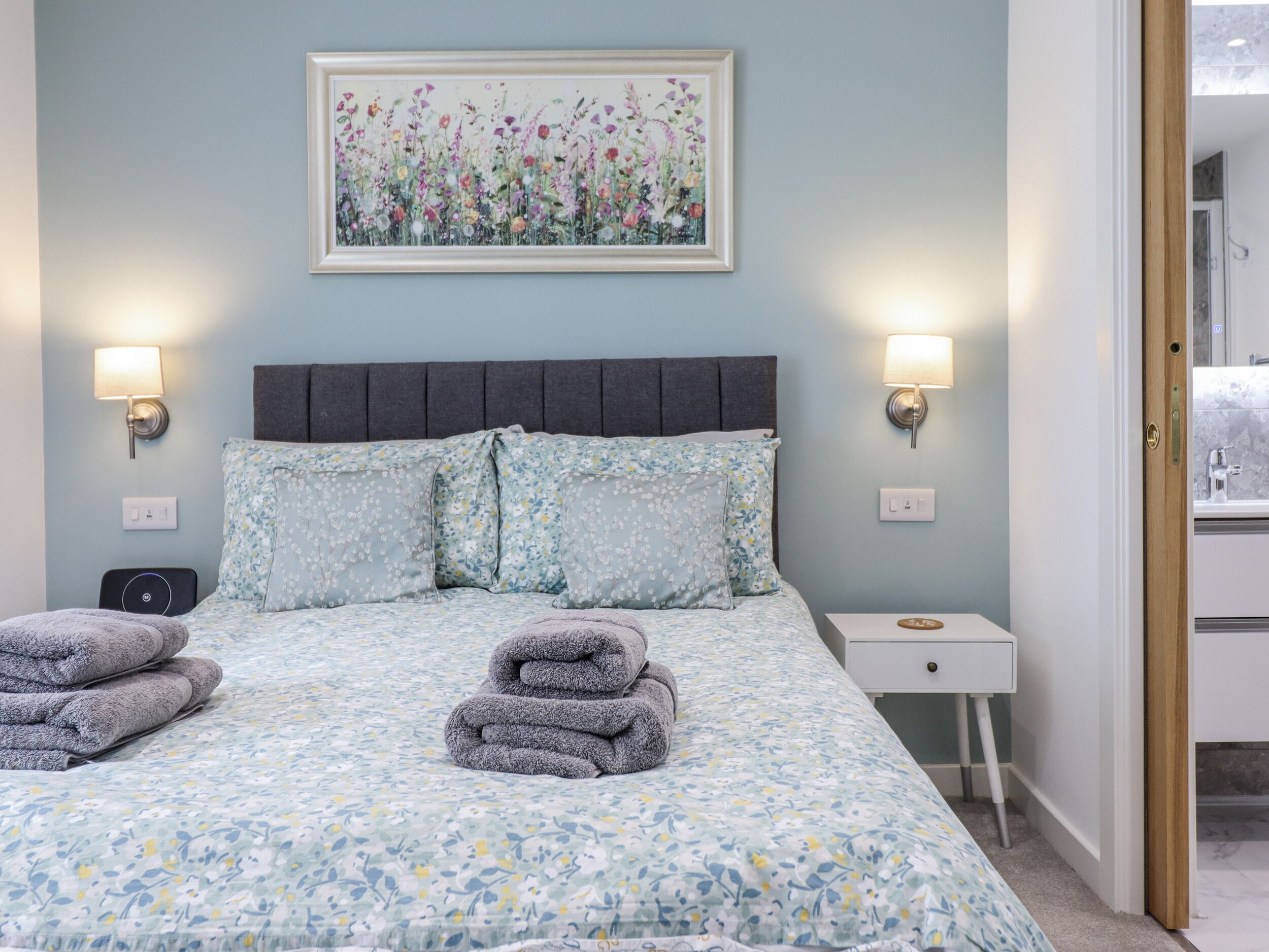 A bedroom with a bed, floral bedding, wall-mounted lamps, a bedside table, and a framed floral painting.