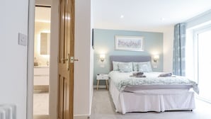 A bedroom with a bed, bedside tables, a nightstand, a window with curtains, and a door leading to a bathroom. - Neptune Cottage (Conwy)