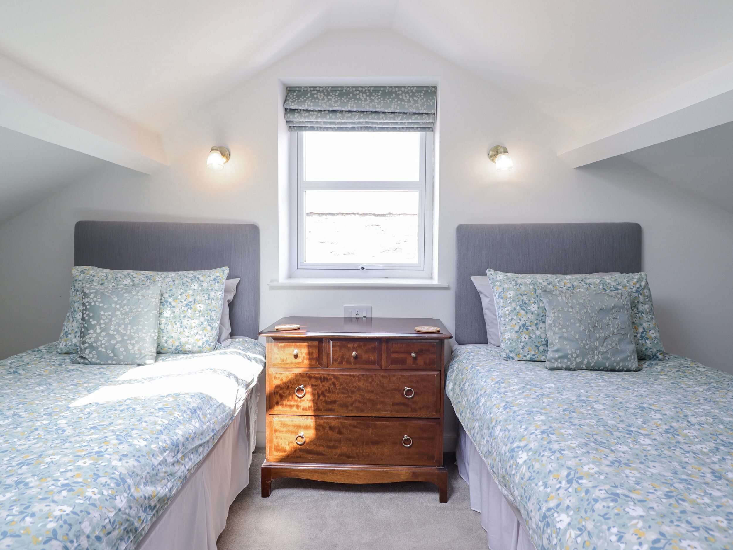 A bedroom with two single beds, a wooden dresser, a window with a view, and two wall-mounted lights.
