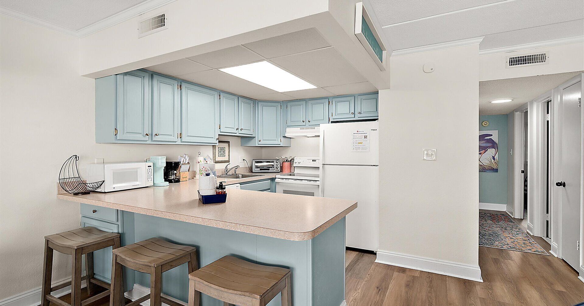 Une cuisine avec des meubles de couleur bleu clair, un réfrigérateur blanc, un four à micro-ondes et un grille-pain. On y trouve trois tabourets en bois et un petit îlot central avec un évier.