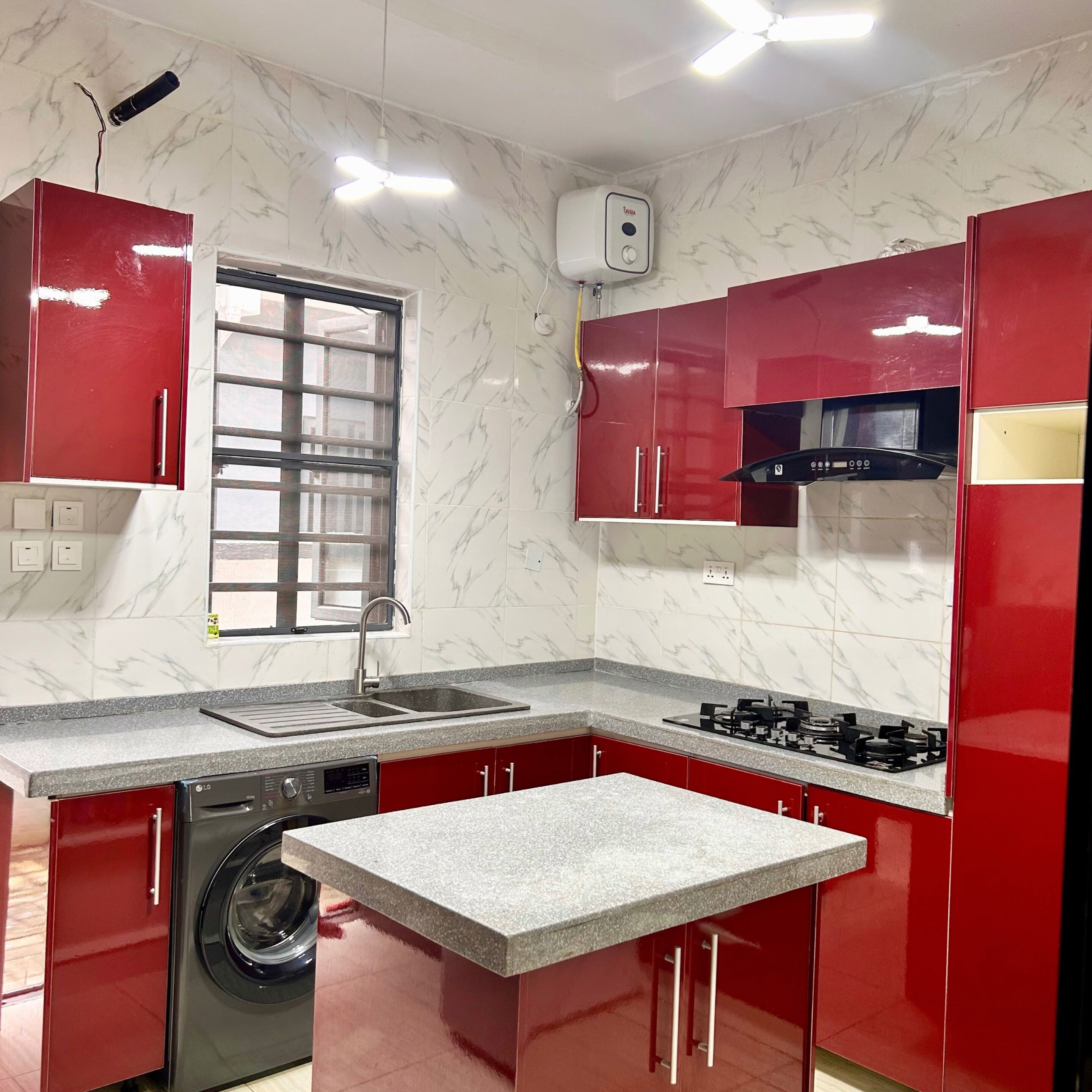 A compact kitchen with red cabinets, a built-in washing machine, a gas stove, and a window with a metal grill.