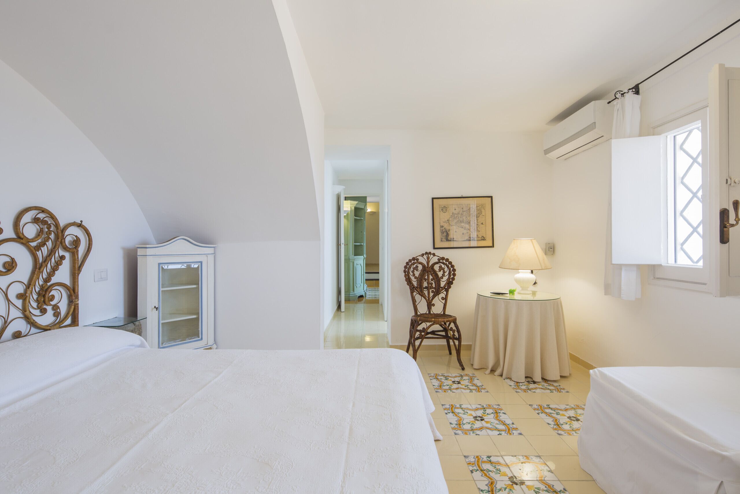 A bedroom with two beds, a cabinet, a chair, a table with a lamp, and a window with curtains.