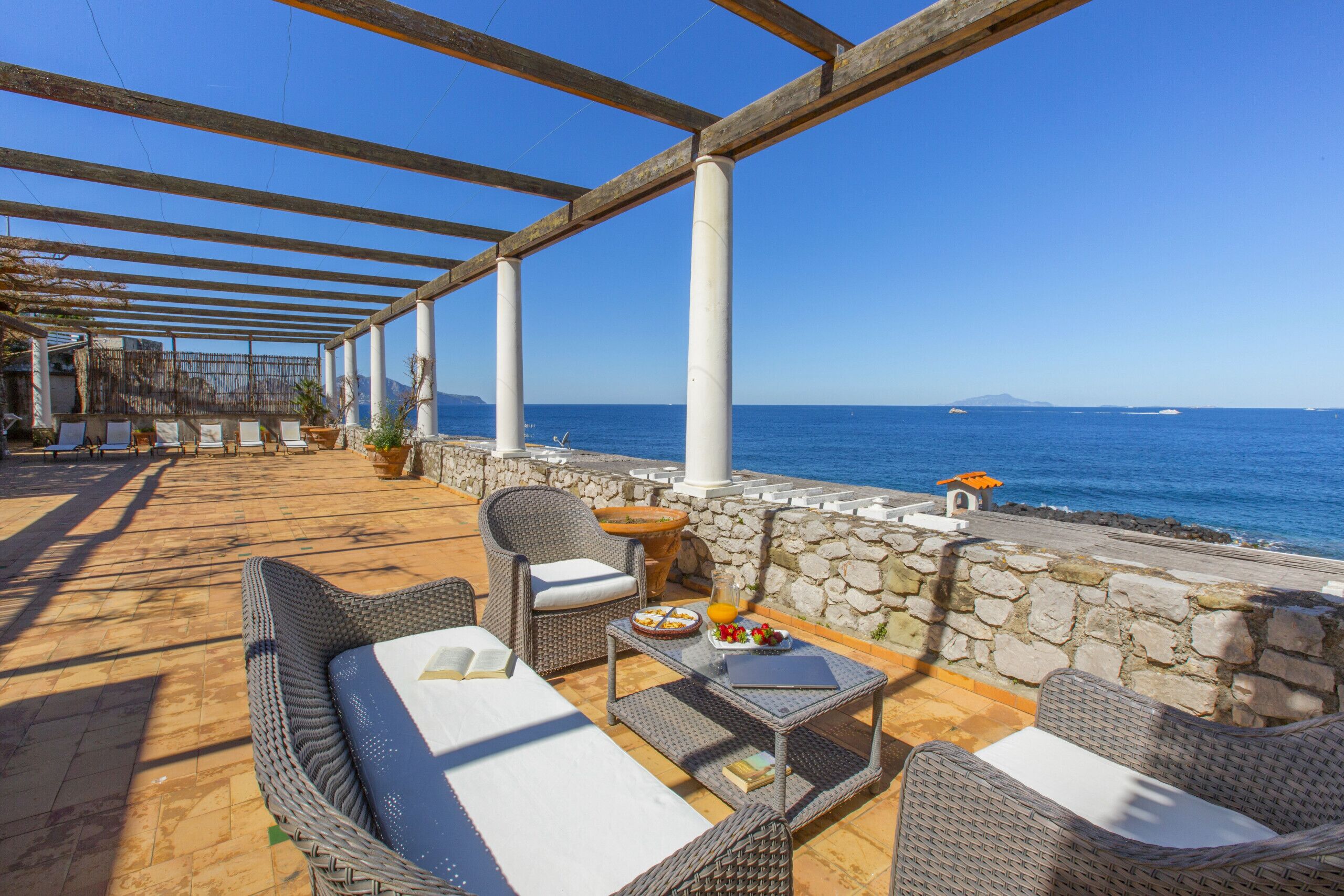 A covered outdoor seating area with wicker furniture, a stone wall, and a view of the sea.
